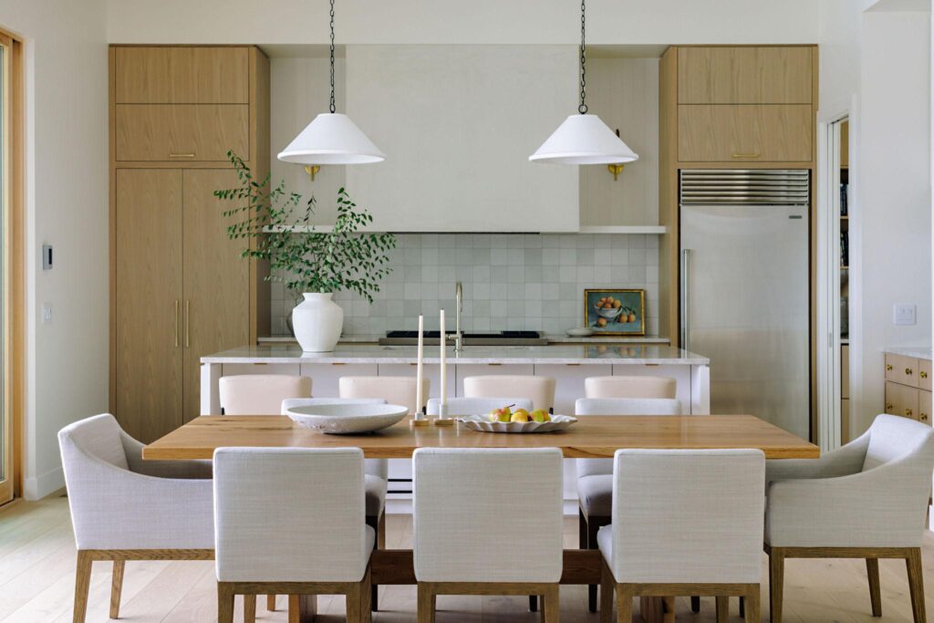 Framed doorway from the main kitchen looking into the butler's pantry, showing floor-to-ceiling white oak cabinets with round brass knobs, zellige tile backsplash, marble countertop, panel-ready dishwasher and wide-plank oak flooring, white cabinetry and tile backsplash visible in the foreground kitchen