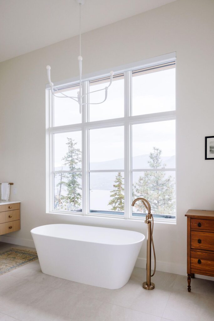 Freestanding soaker tub with brass filler beneath tall windows overlooking Okanagan Lake